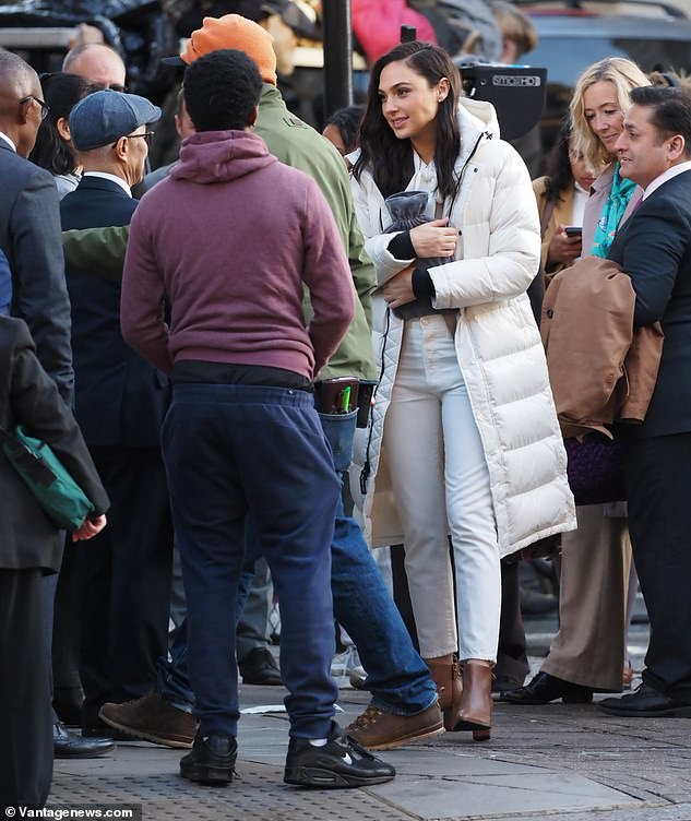 Gal Gadot shrouds herself in a padded coat and hugs a hot water bottle as attempts to keep warm while filming an advert in London